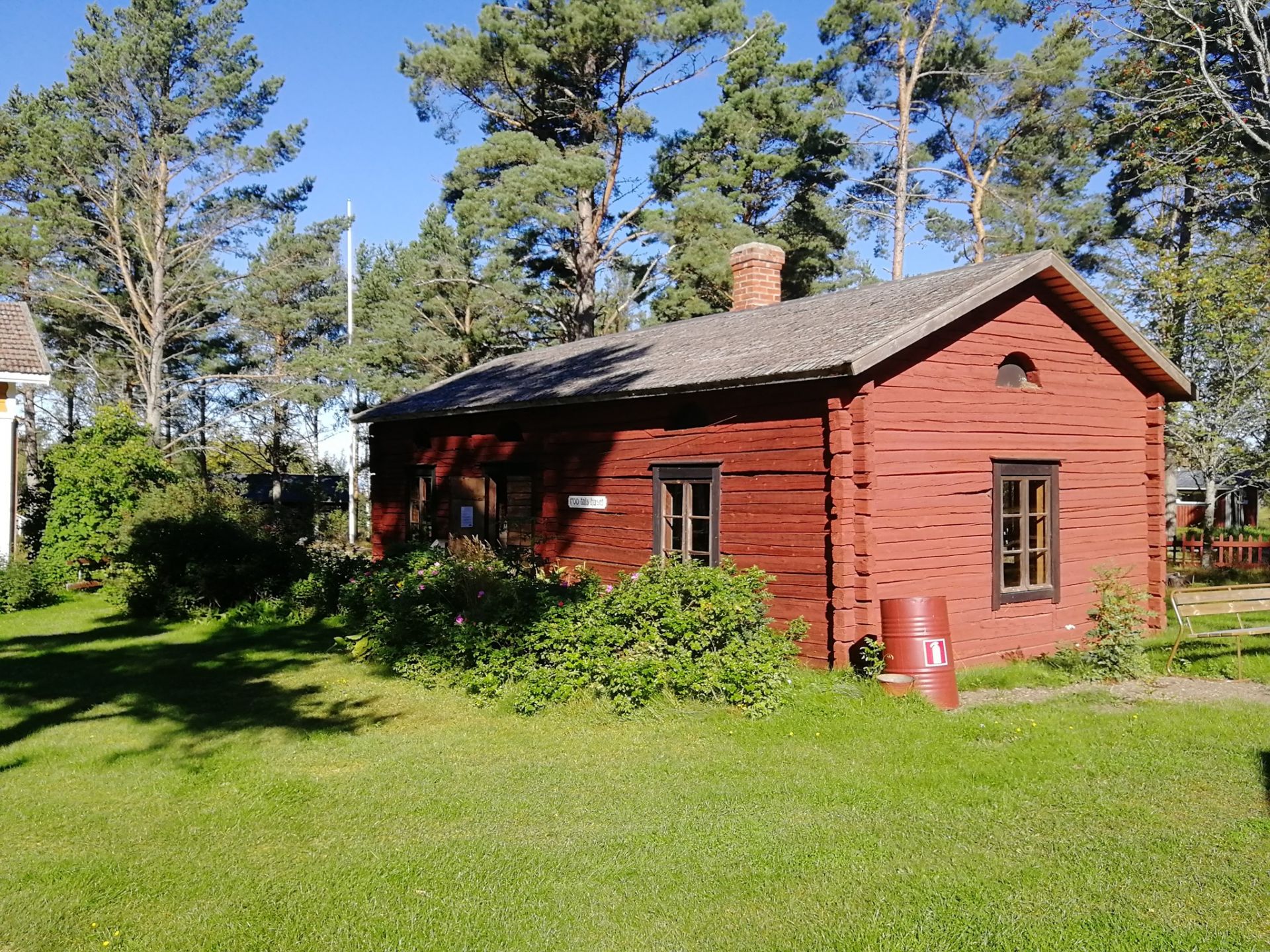 Röd timmerstuga från 1700-talet, omgiven av grönska och med blå himmel.