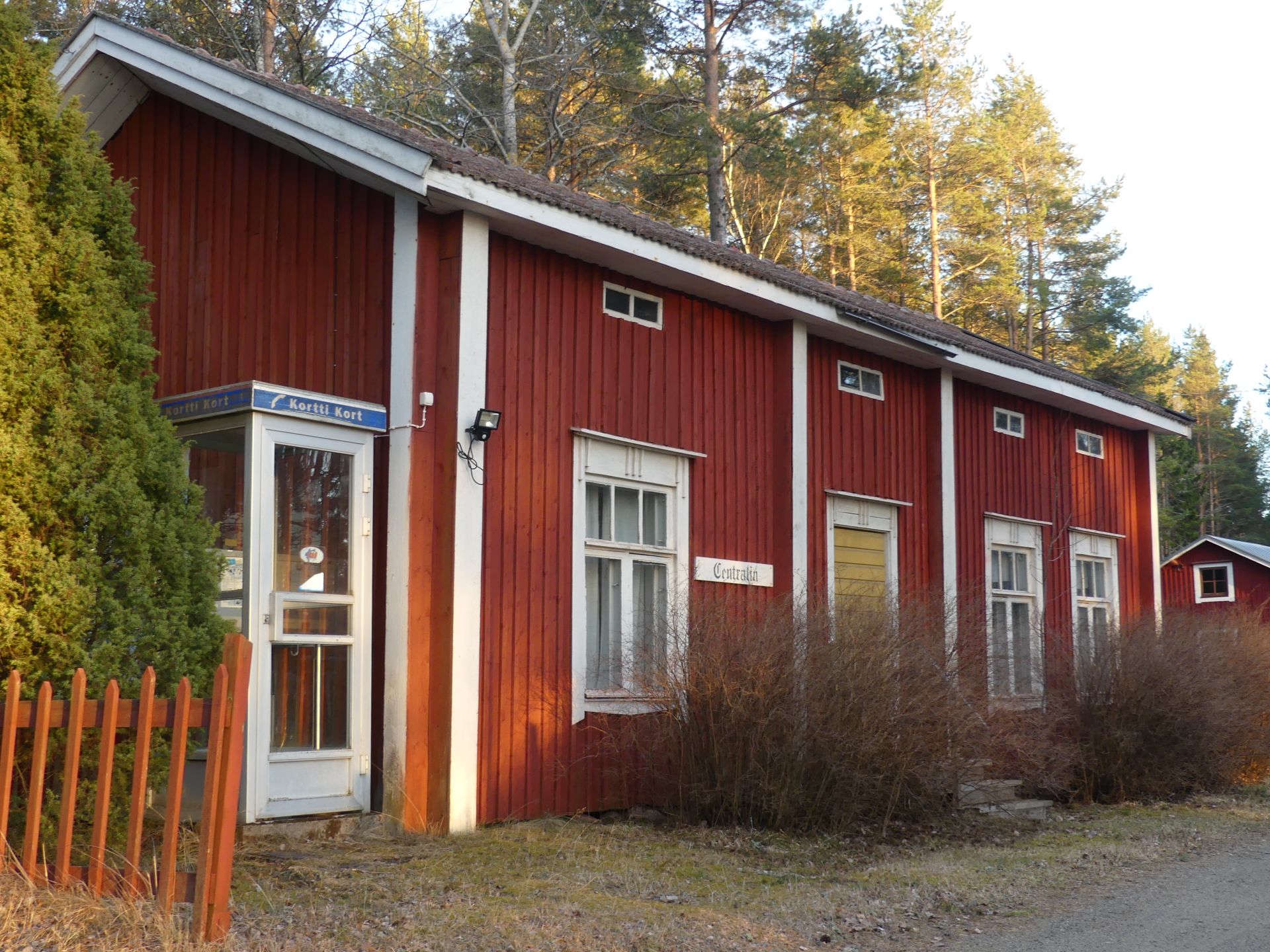 Röd byggnad med vit dörr och skyltar för Centralin och telefonkiosk.