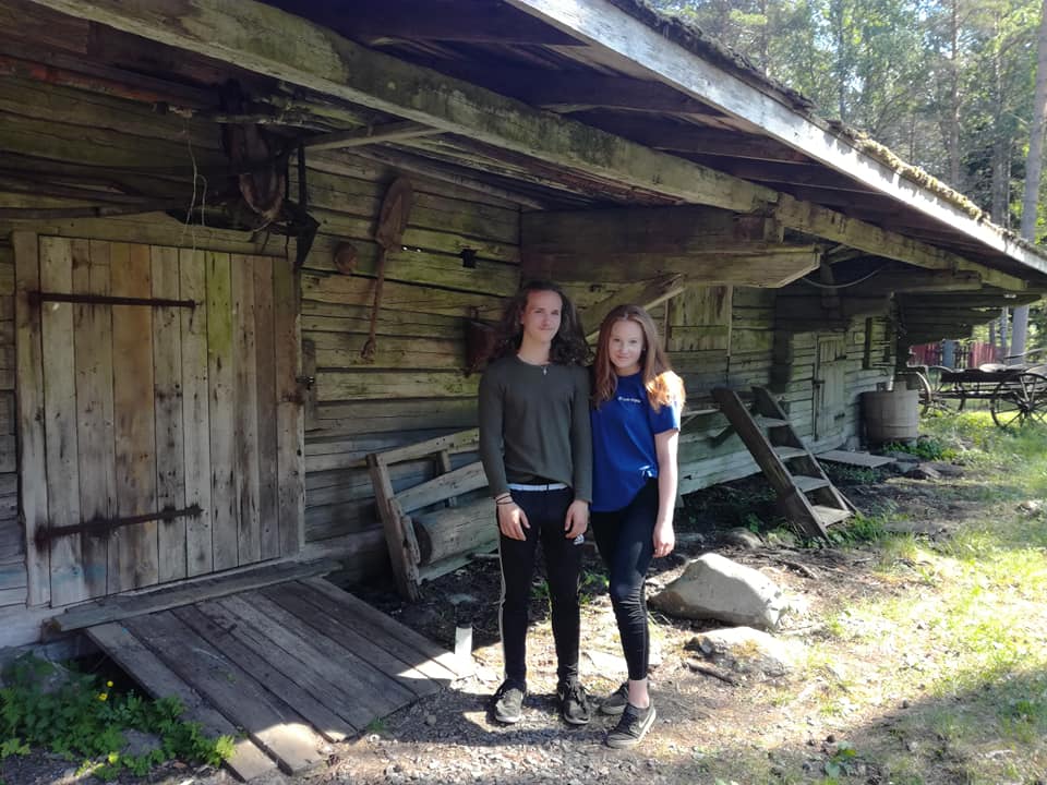 Två ungdomar framför en gammal träbyggnad med verktyg hängande på väggen.