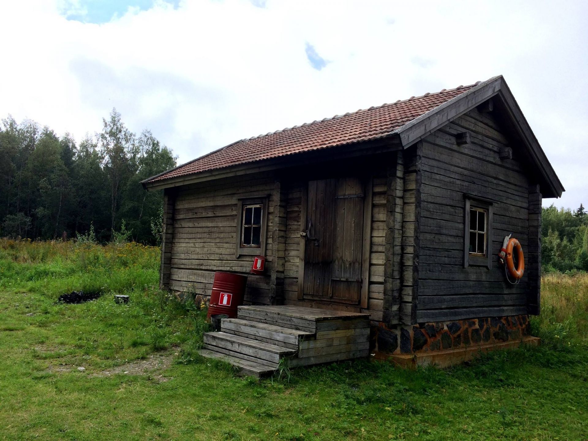 Grå rökbastu i timmer med brandsläckare och livboj, belägen på gräsmark nära skog.