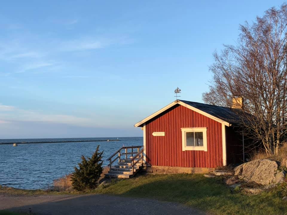 Rött strandnära trähus med trappa ner mot havet under klarblå himmel.