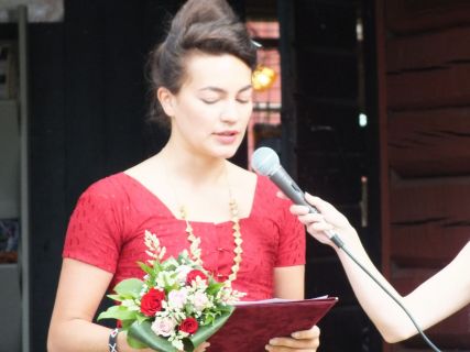 Punaiseen mekkoon pukeutunut nainen pitää juhlapuhetta Kilenin tapahtumassa, kädessään kukkakimppu ja mikrofoni.